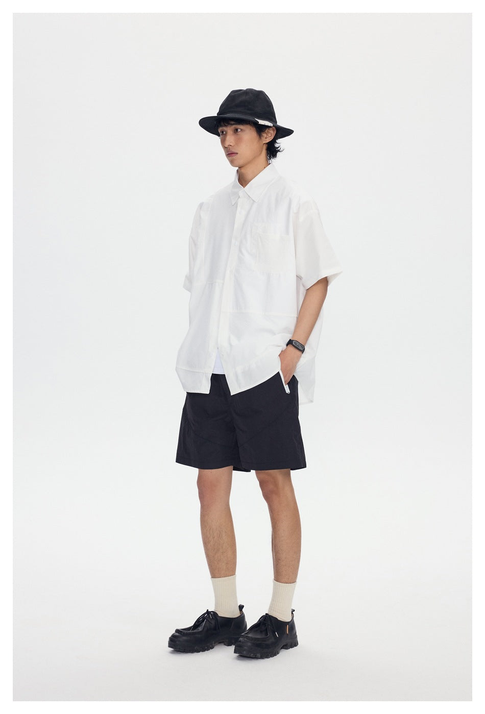 Model wearing a white short-sleeve button-up shirt and black shorts, with a black hat and shoes.