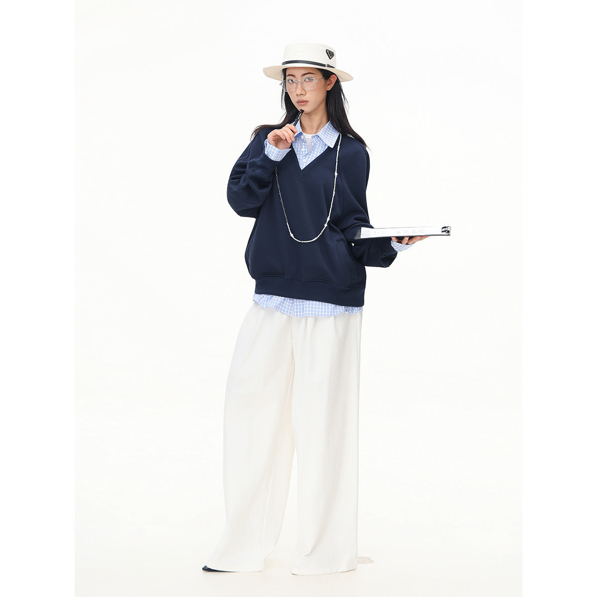 Full-length model in a navy V-neck sweatshirt, blue checked shirt, wide white trousers, and cream hat.