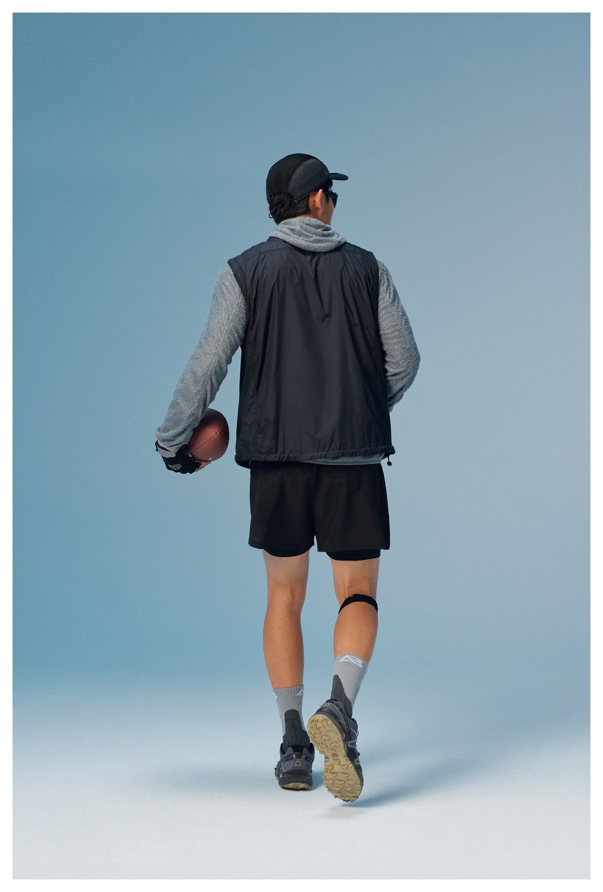 Rear view of a model wearing a black sleeveless vest over a gray fleece top and black shorts.
