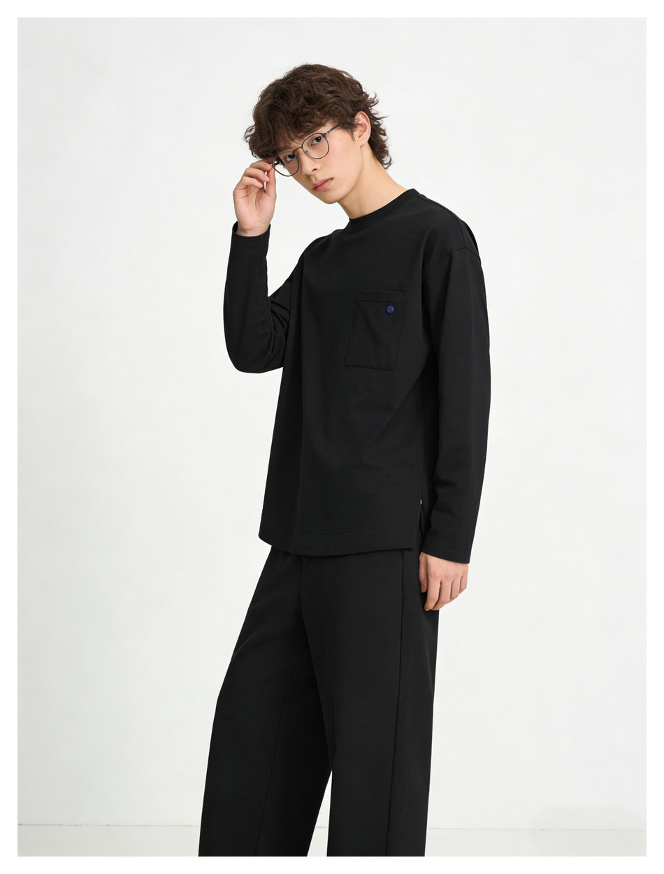 Model in black long-sleeve crew neck top with button-accent chest pocket, hand near glasses, with black trousers