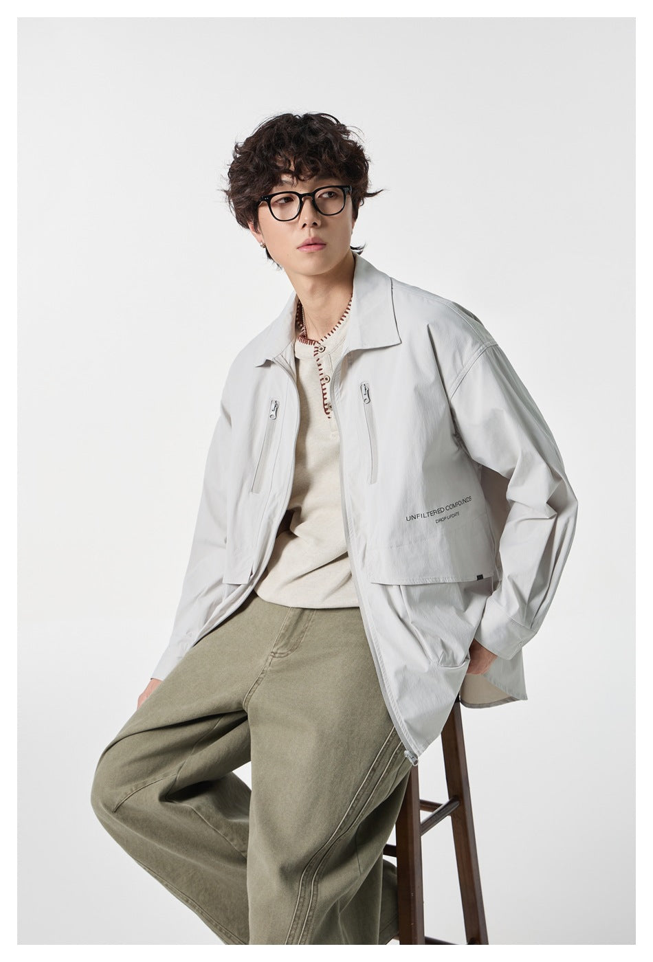 Seated model wearing an open pale gray utility jacket over a cream top with loose olive trousers.