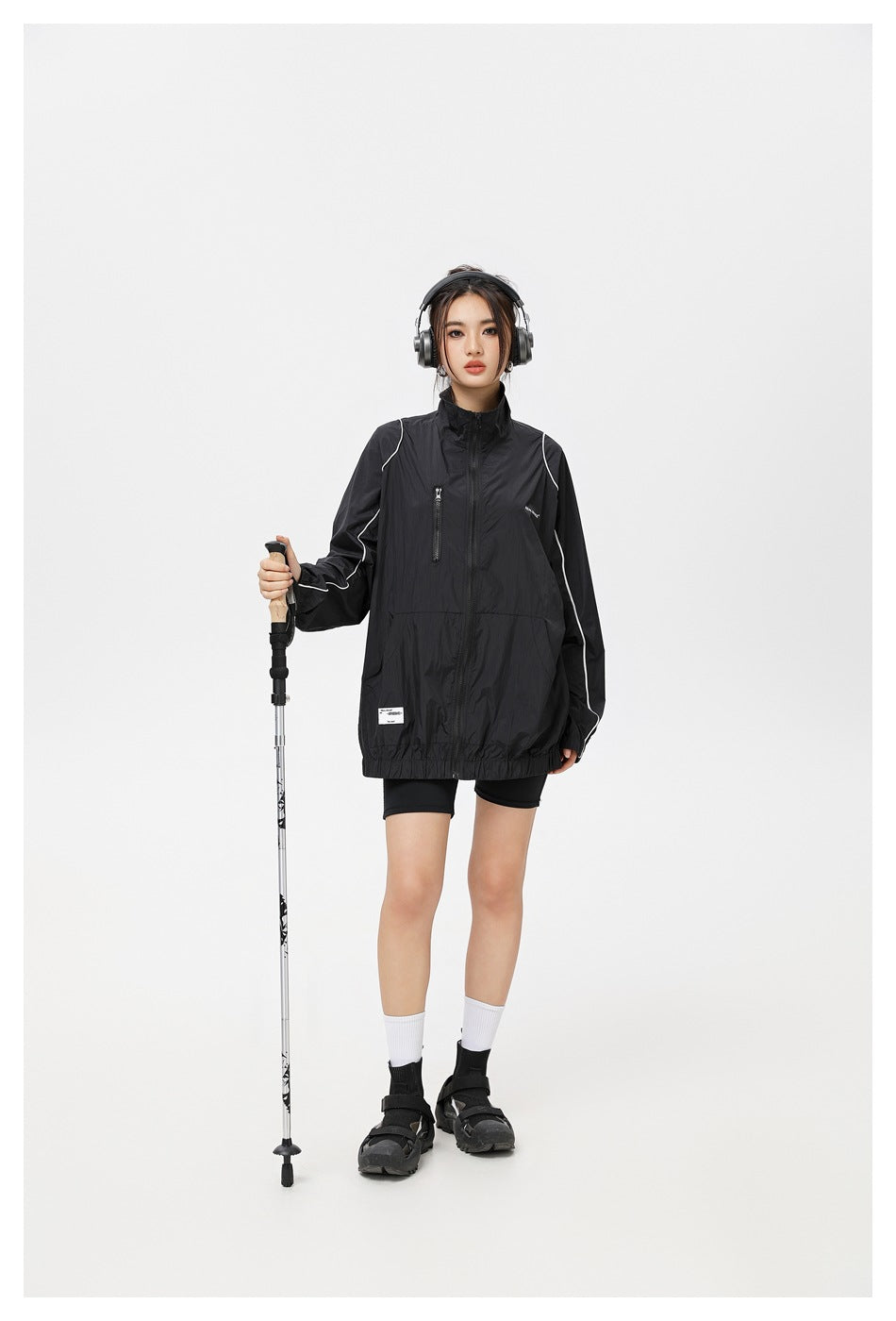 Front view of a model wearing a black zip jacket with white piping, posed beside a trekking pole.