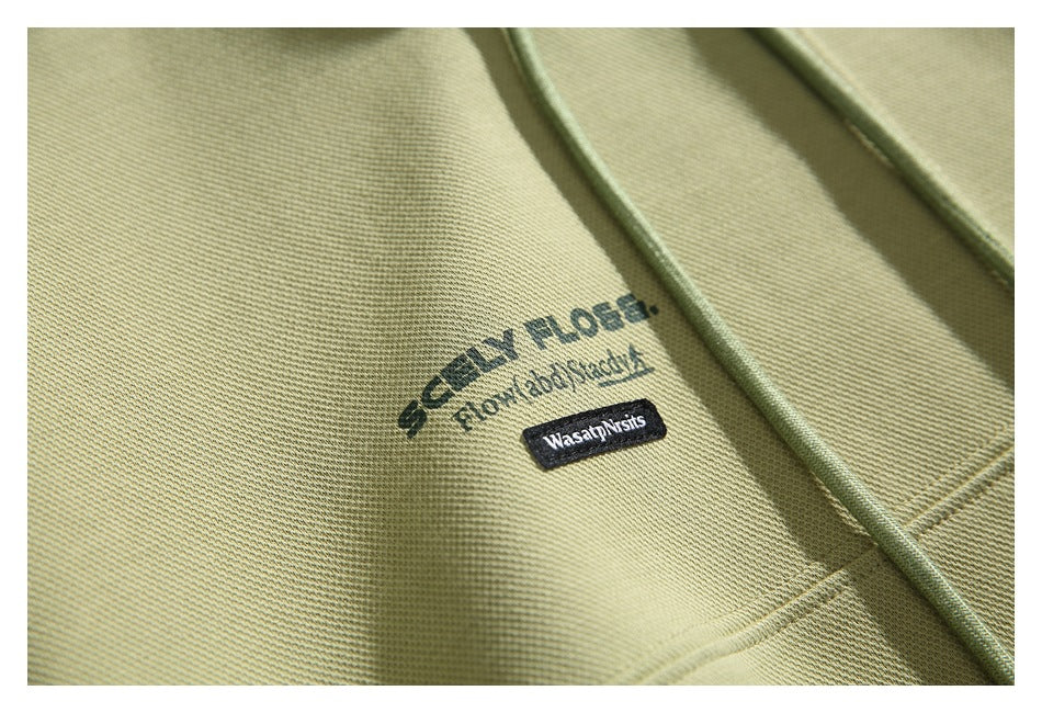Close-up of the olive-green hoodie fabric and small printed chest design beside a drawstring.