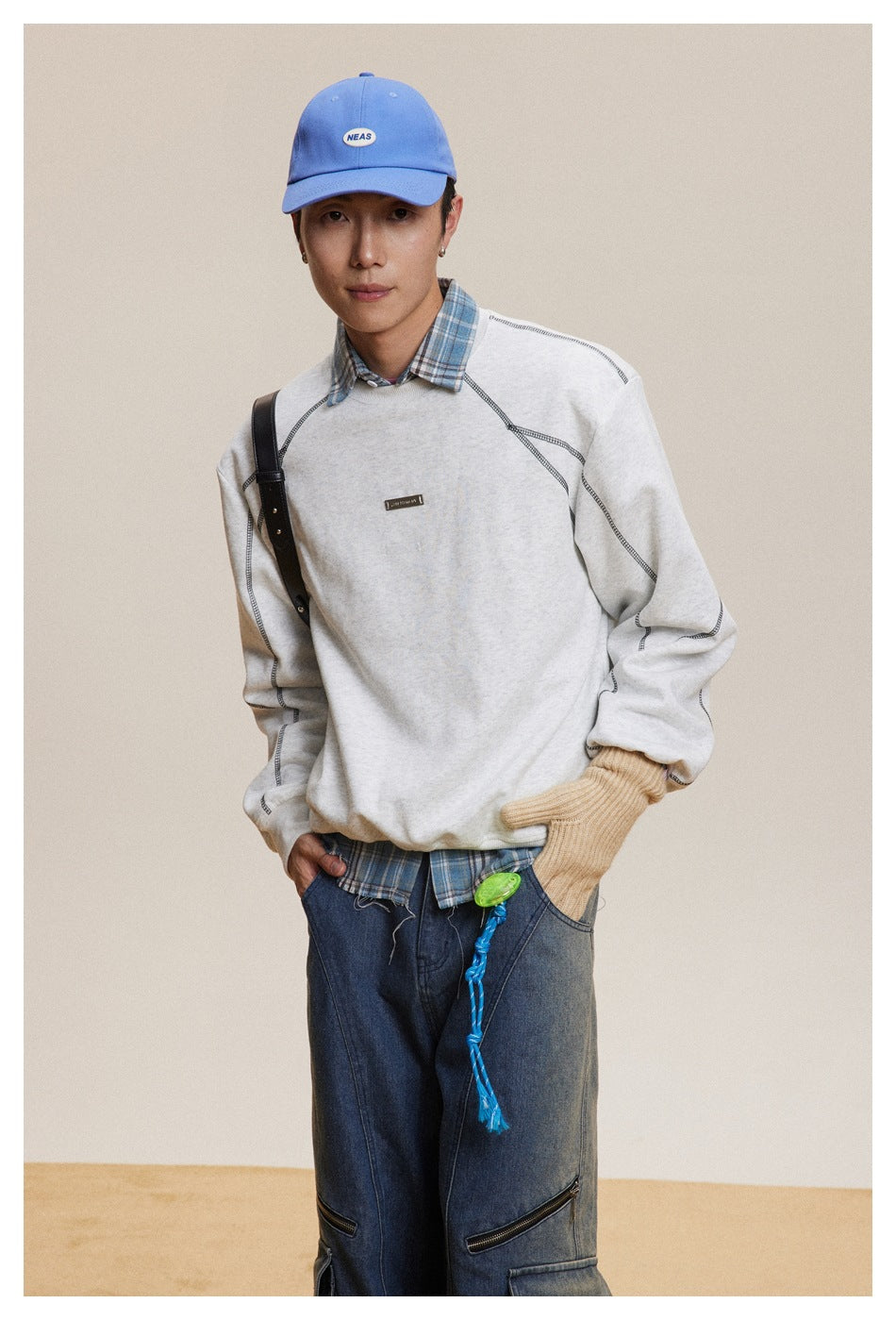 Model wearing a light gray sweatshirt with contrast seam stitching, plaid shirt, blue cap and dark cargo jeans.