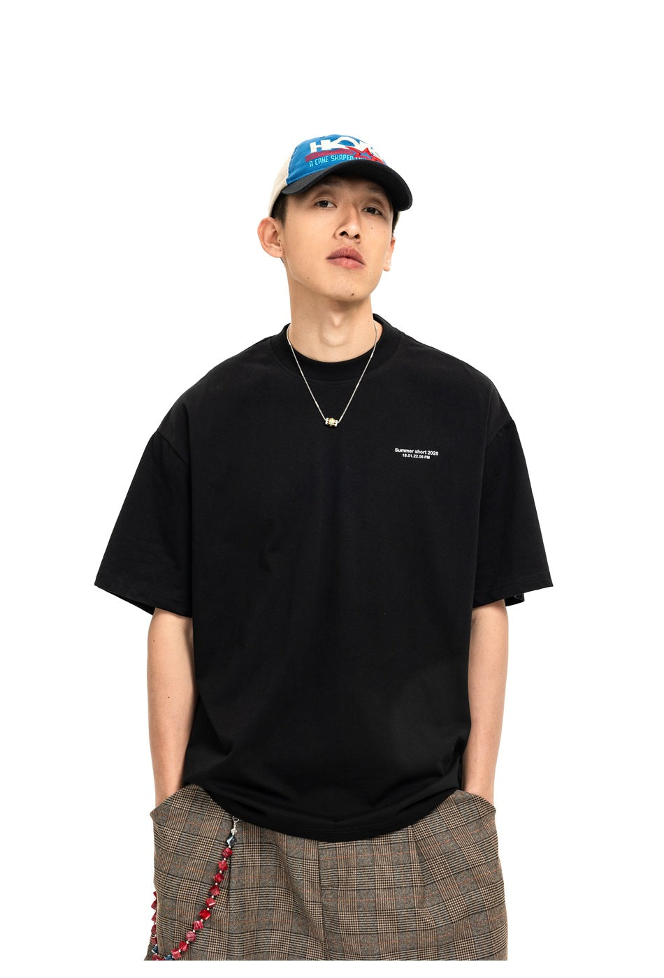 Front view of a model wearing a black T-shirt with small white chest text and brown plaid shorts.