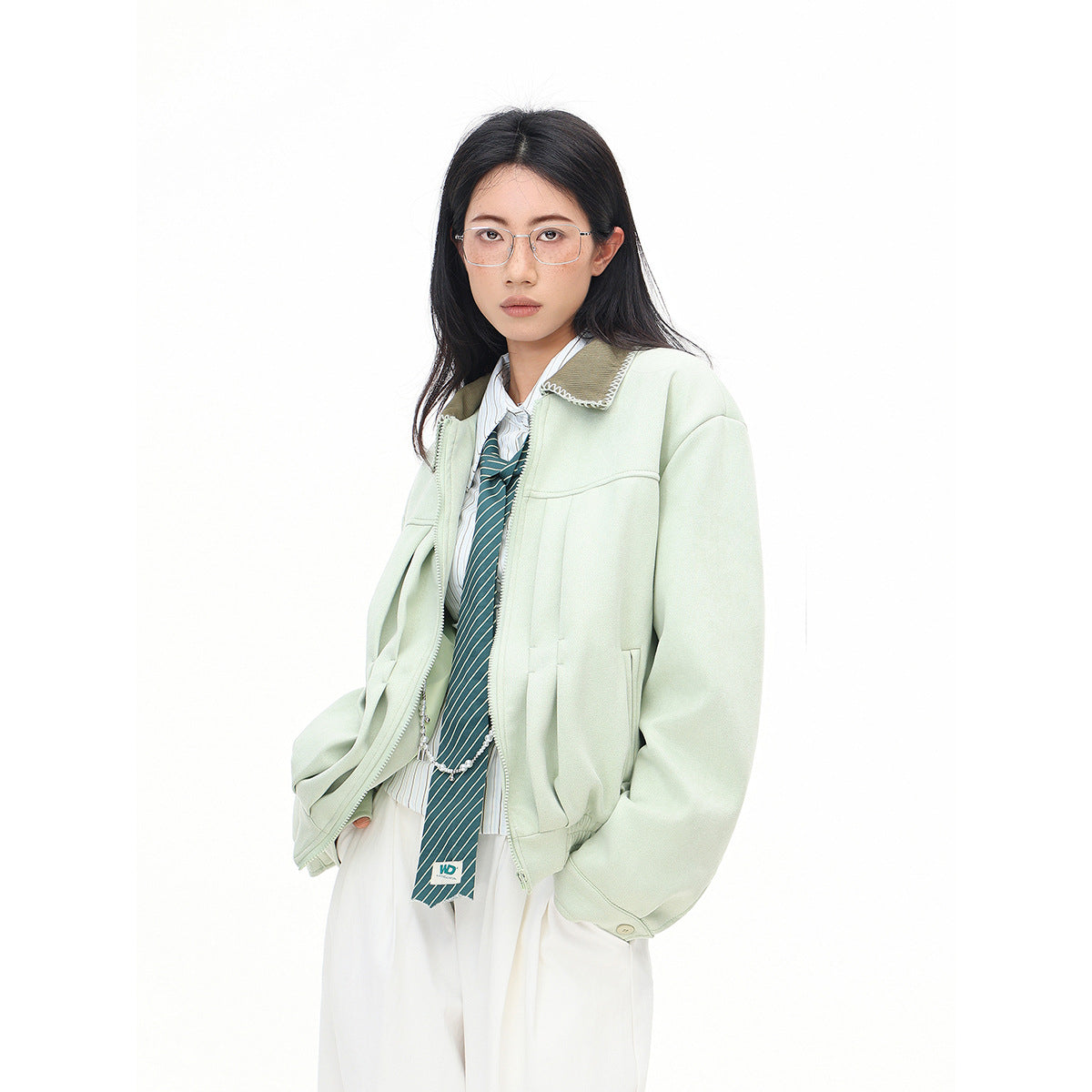 Three-quarter view of a model wearing an open pale mint jacket over a striped shirt and tie with white trousers.