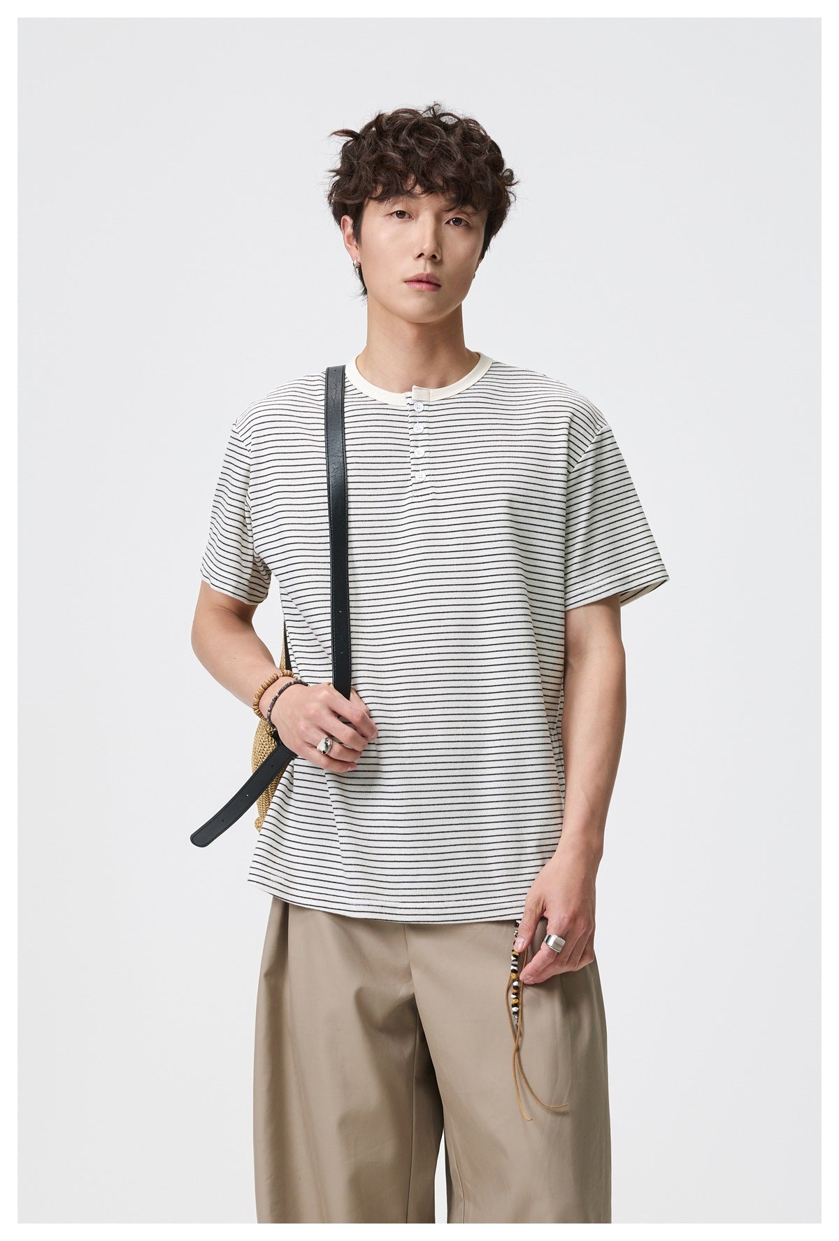 Model wearing an ivory, thin black-striped Henley T-shirt with beige trousers and a dark shoulder strap.