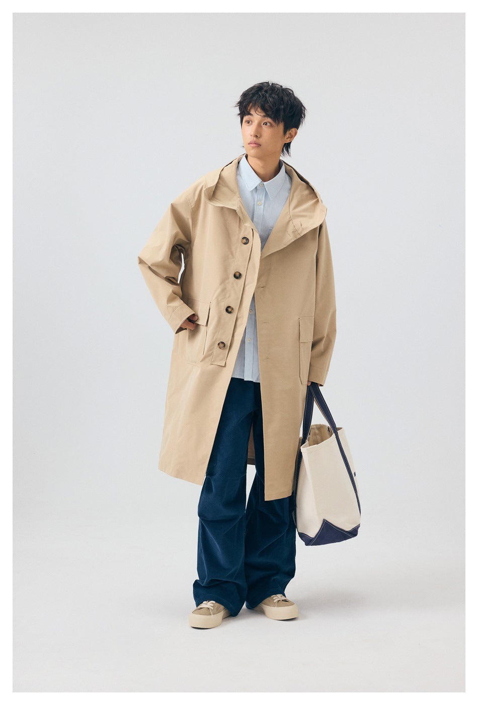 Full-length model wearing a beige hooded coat with navy trousers and carrying a cream tote with navy trim.