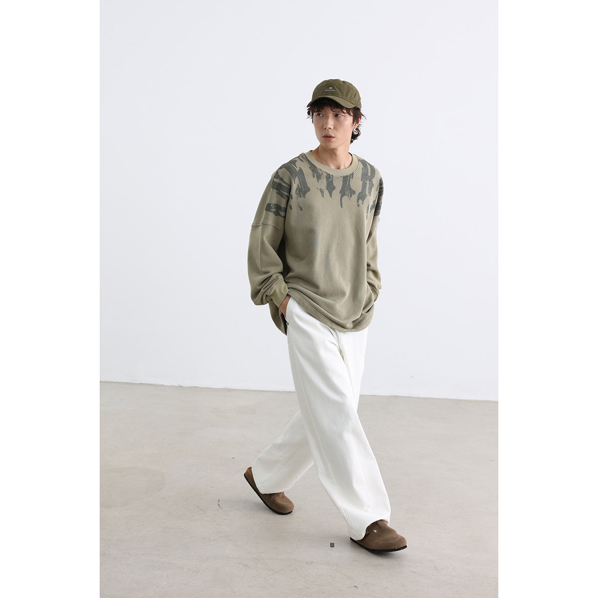 Model wearing an oversized khaki sweatshirt with dark abstract shoulder graphics, white wide-leg pants and an olive cap.