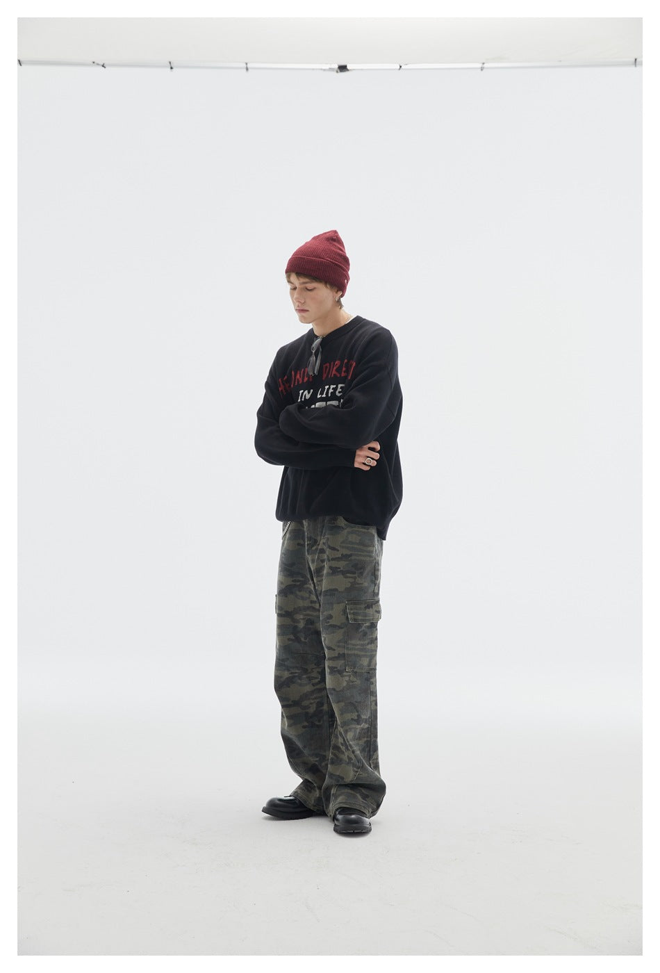 Model with folded arms wearing a black lettered crewneck sweater, burgundy beanie, and camouflage cargo pants.