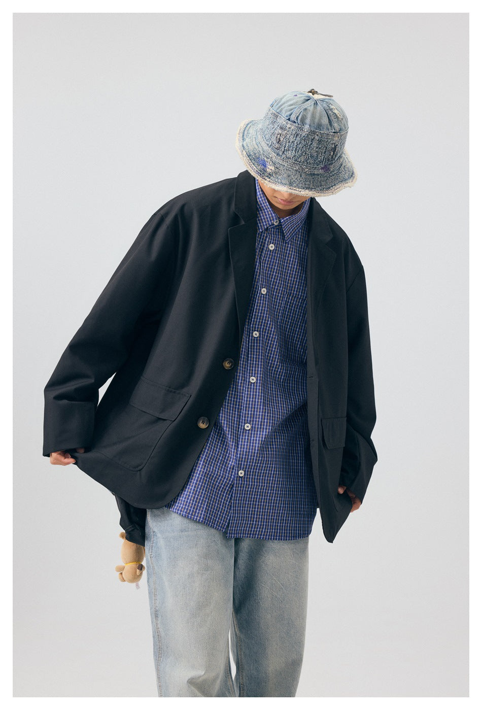Model wearing an oversized black blazer over a blue checked shirt and light jeans, looking down beneath a denim bucket hat.