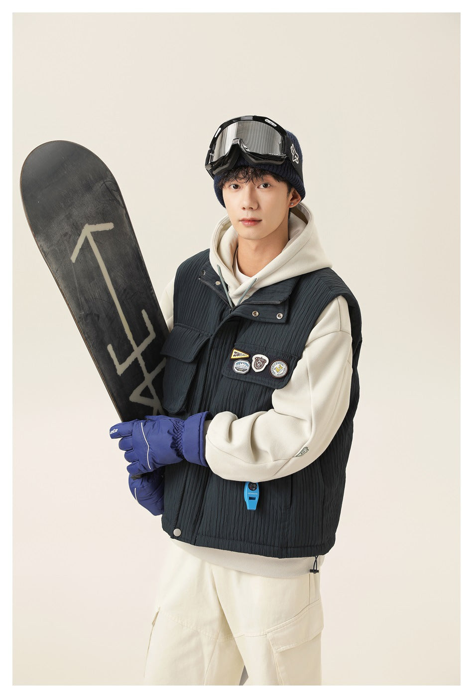Model wearing a dark charcoal quilted utility vest over an ivory hoodie, holding a snowboard.