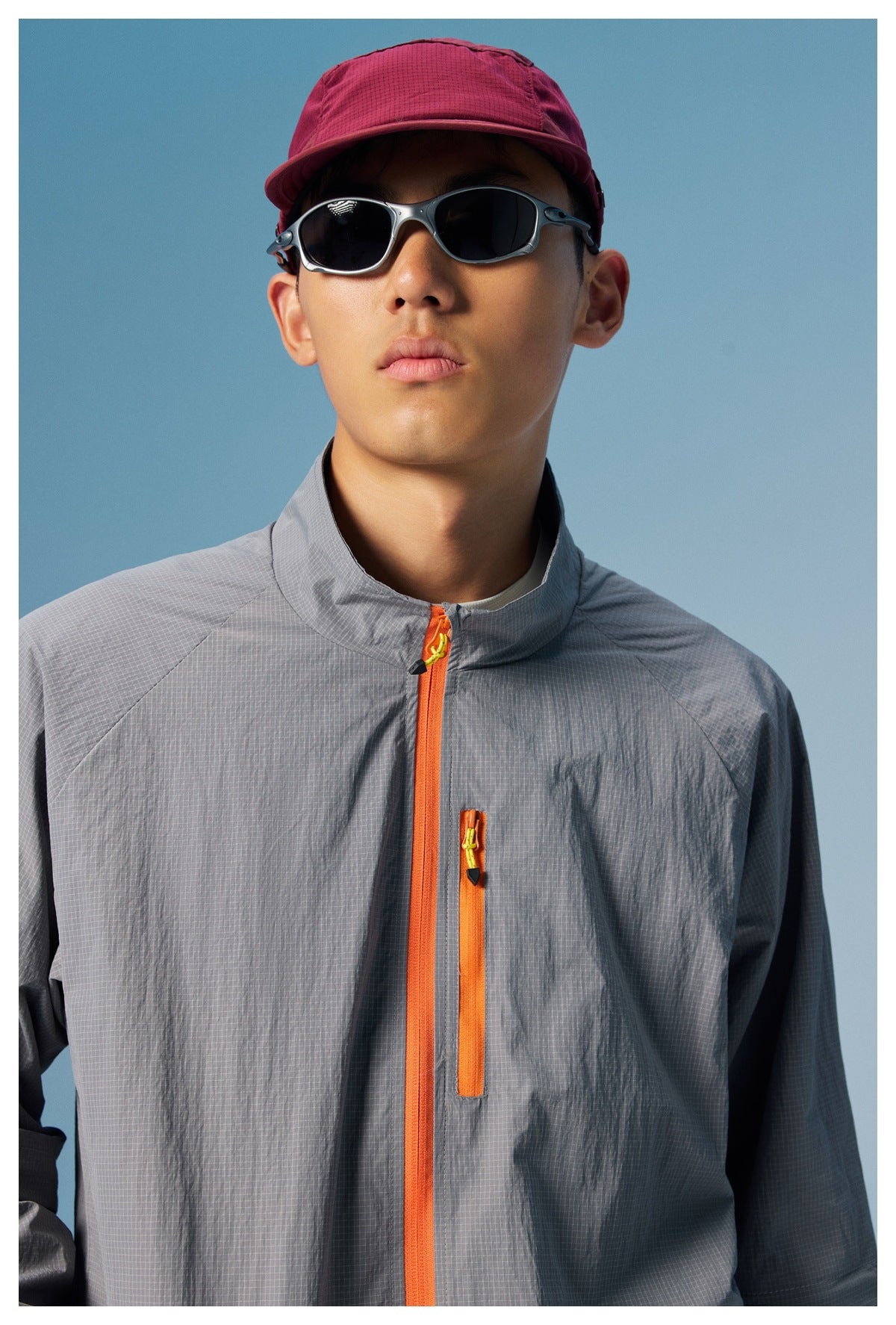 Model wearing a gray grid-textured zip jacket with bright orange zippers, burgundy cap, and dark sunglasses.