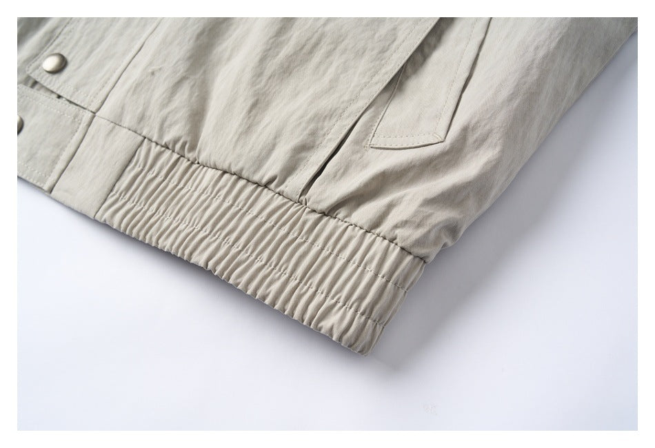 Close-up of a light beige jacket's gathered elastic hem, pocket opening and metal snap.