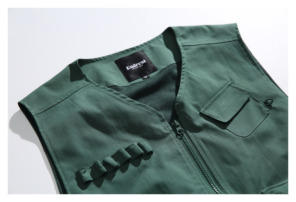 Close-up of a dark green utility vest showing the zipper, chest pocket, webbing loops, mesh lining, and sewn label.