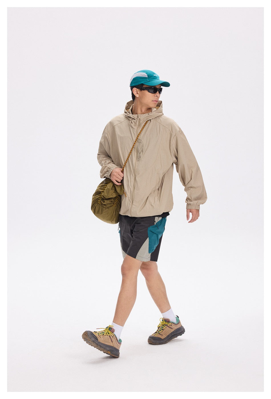 Full-length model wearing a beige hooded jacket, color-block shorts, teal cap and hiking shoes.