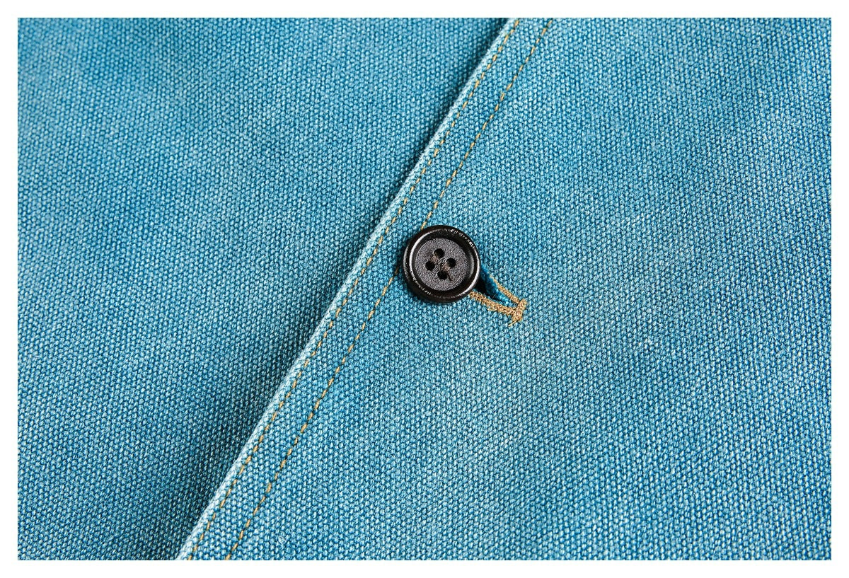Close-up of blue woven fabric with a dark button, ochre buttonhole stitching, and diagonal seam.