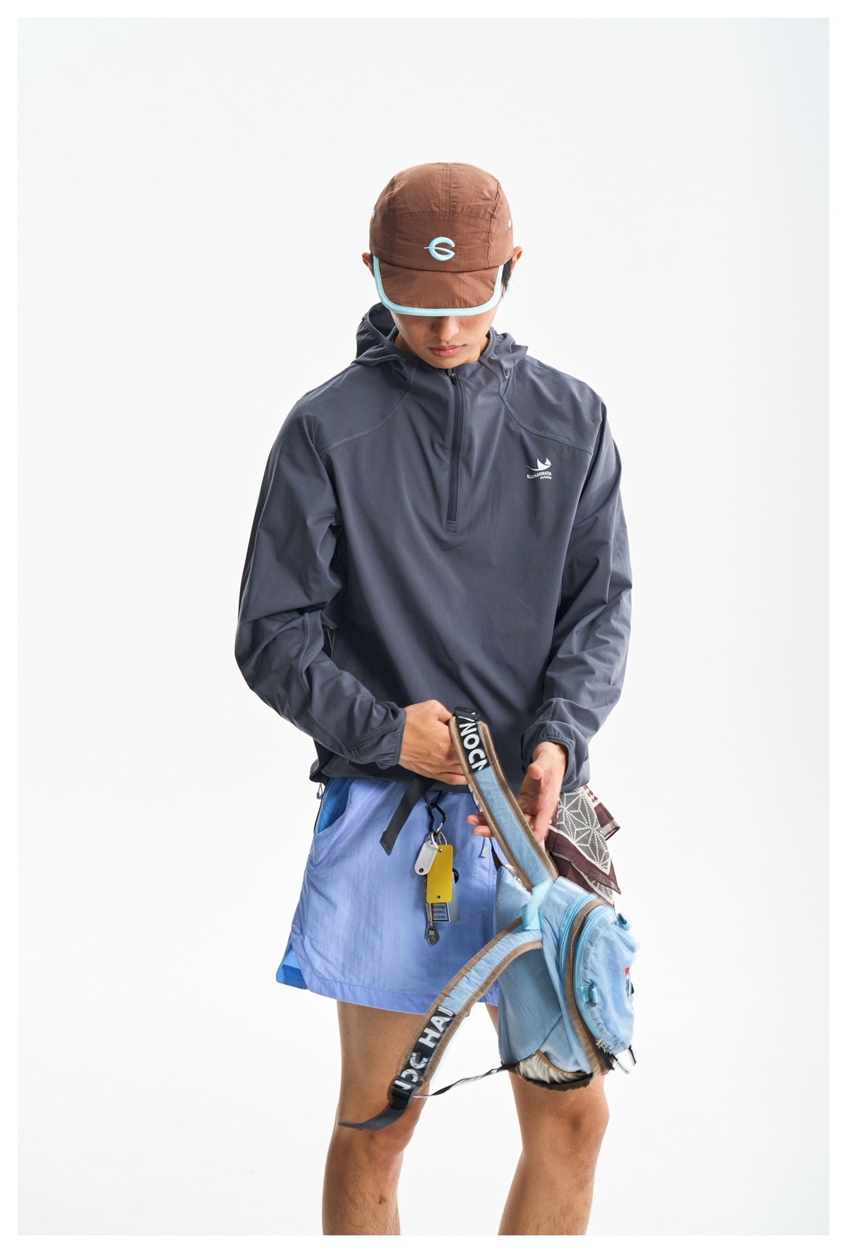 Model wearing a charcoal quarter-zip hooded pullover with light blue shorts and a brown cap.