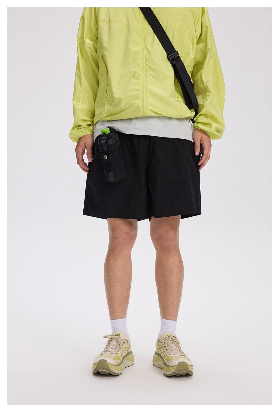 Cropped model wearing black knee-length shorts with an attached utility pouch, paired with a lime jacket and beige sneakers.