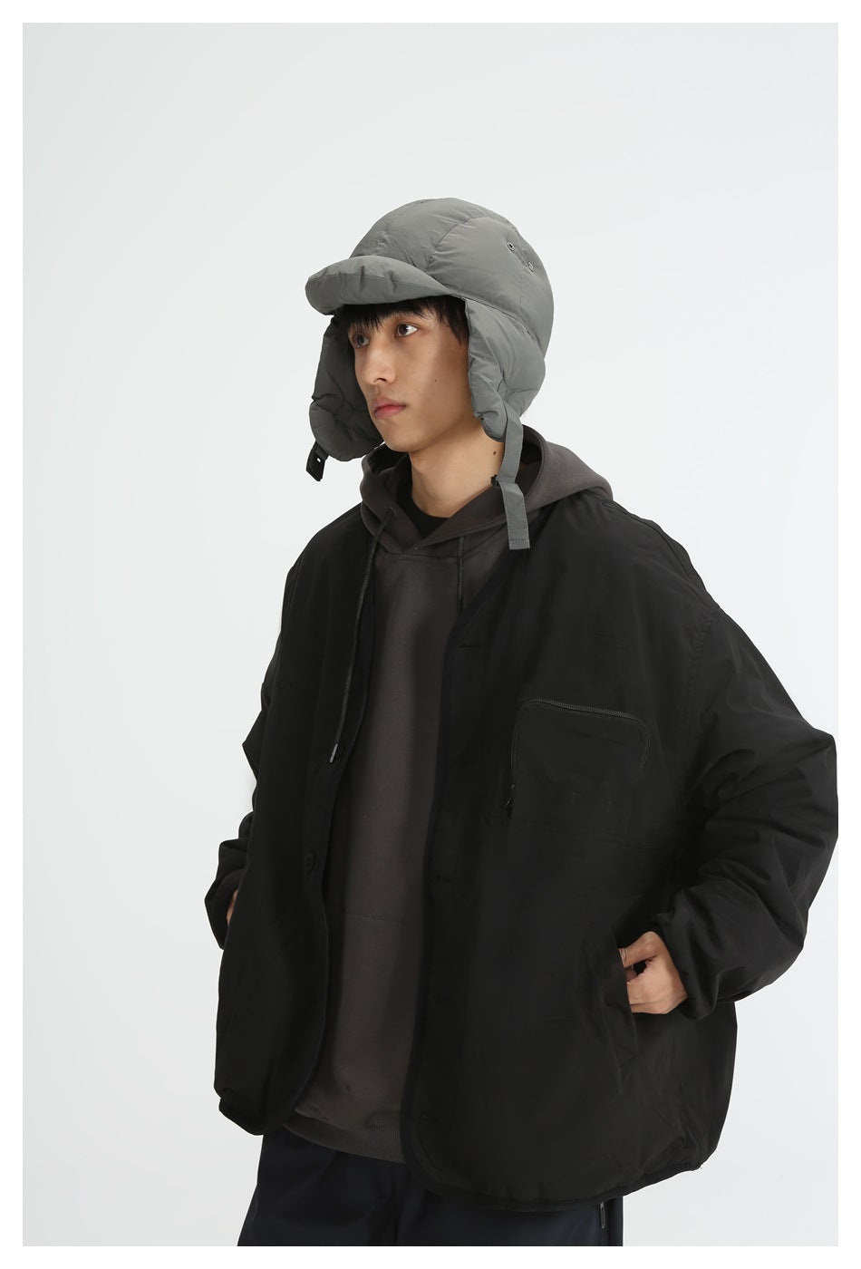 Close-up of a model in a black collarless jacket with zip chest pocket over a hoodie, wearing a gray padded cap