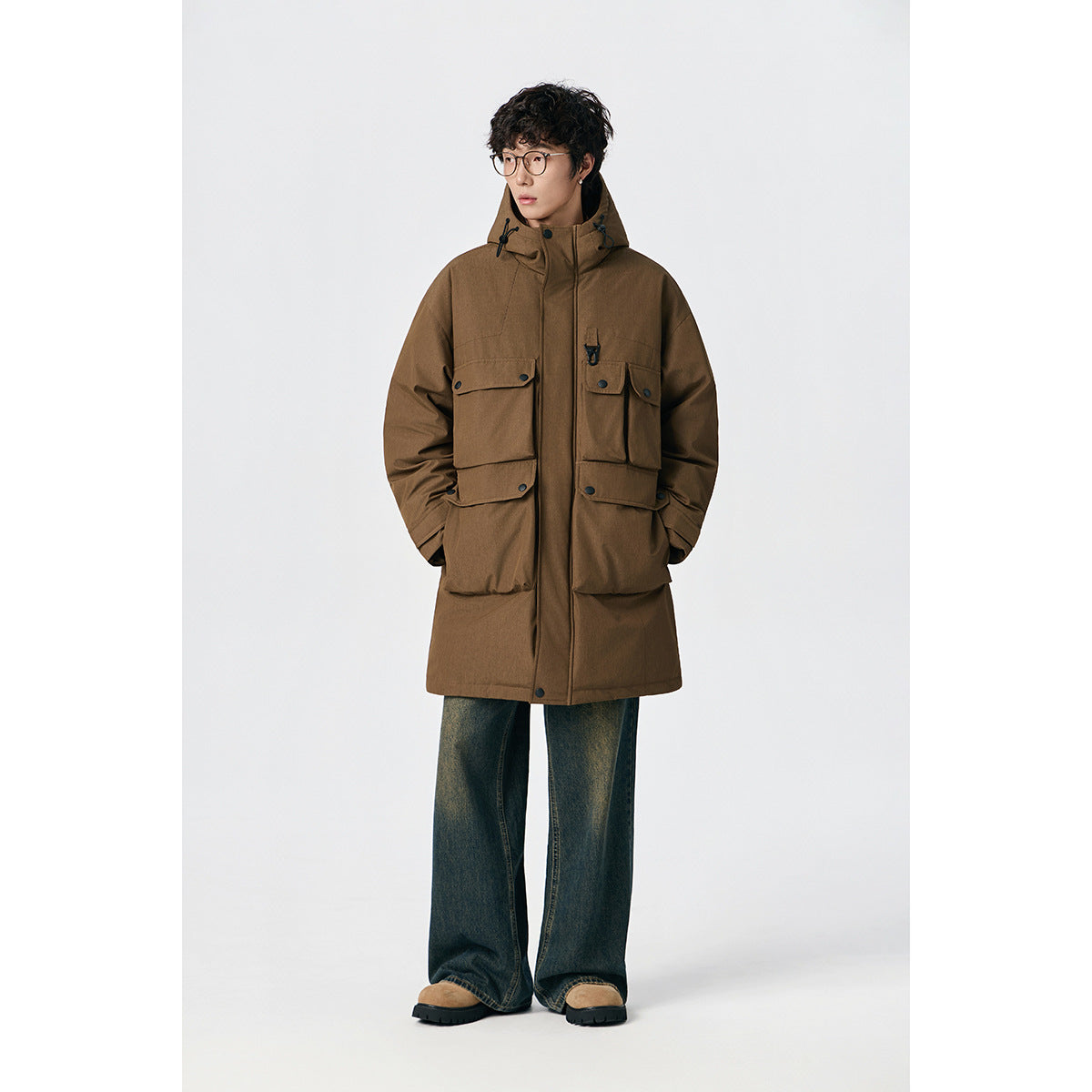 Model wearing a brown hooded long coat with multiple flap pockets, dark wide-leg jeans and tan shoes.