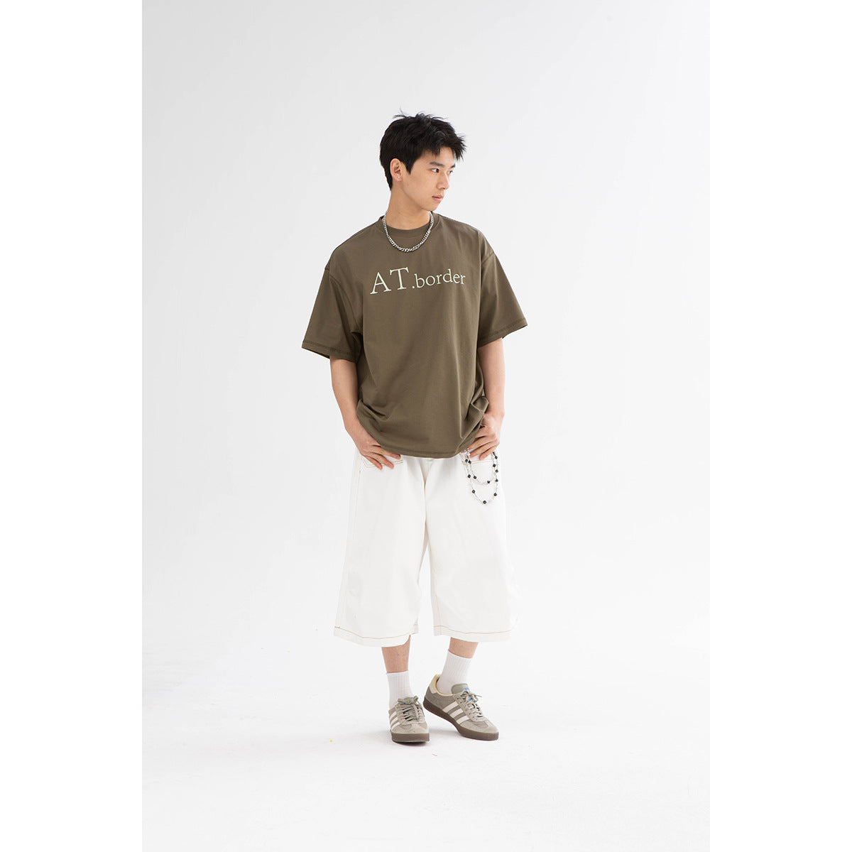Model wearing an olive-brown graphic T-shirt and white wide-leg shorts.