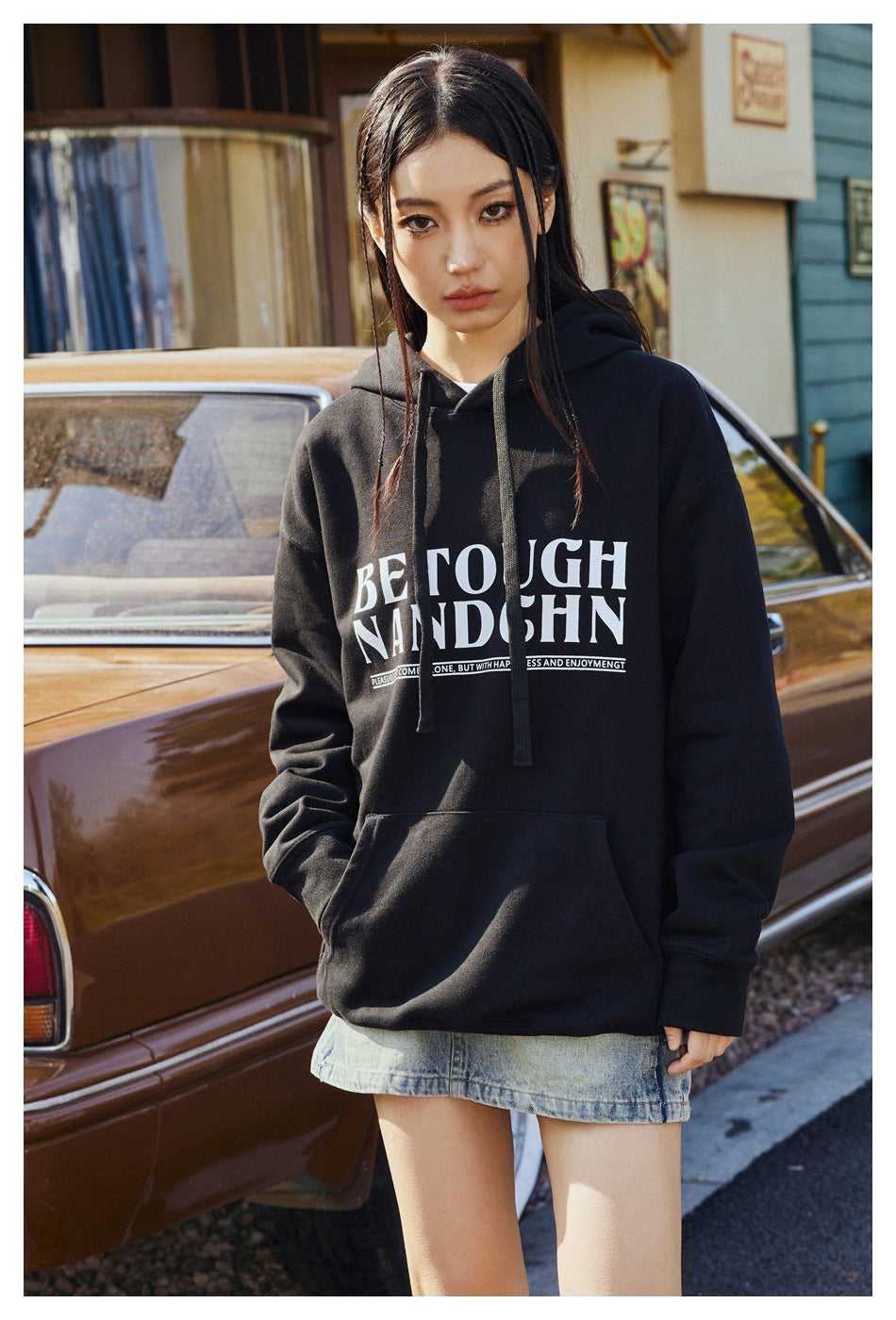 Model wearing a black pullover hoodie with drawstrings, a kangaroo pocket, and large white chest lettering.