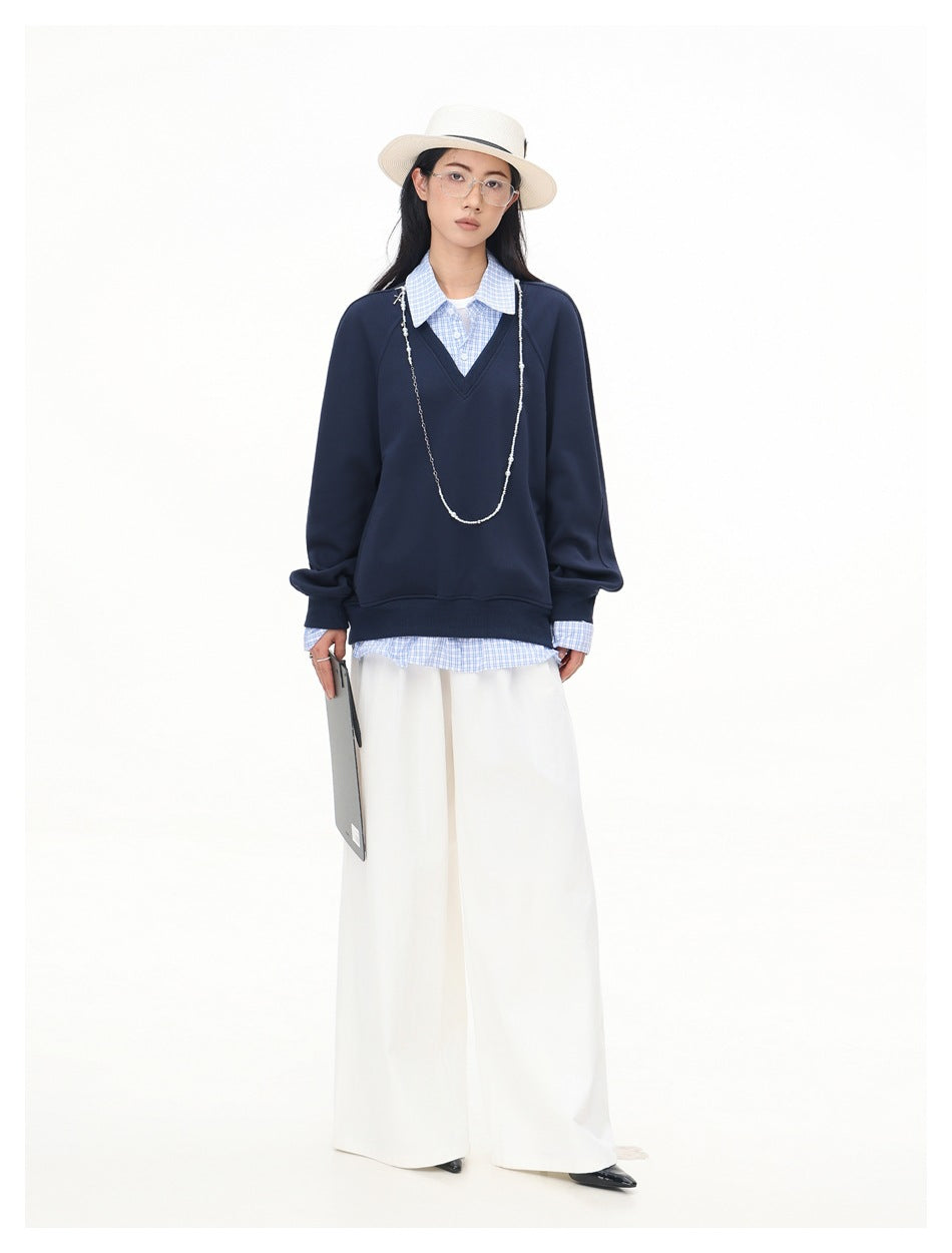 Model wearing navy V-neck sweatshirt with blue checked shirt layer, white wide-leg pants, straw hat and long necklace