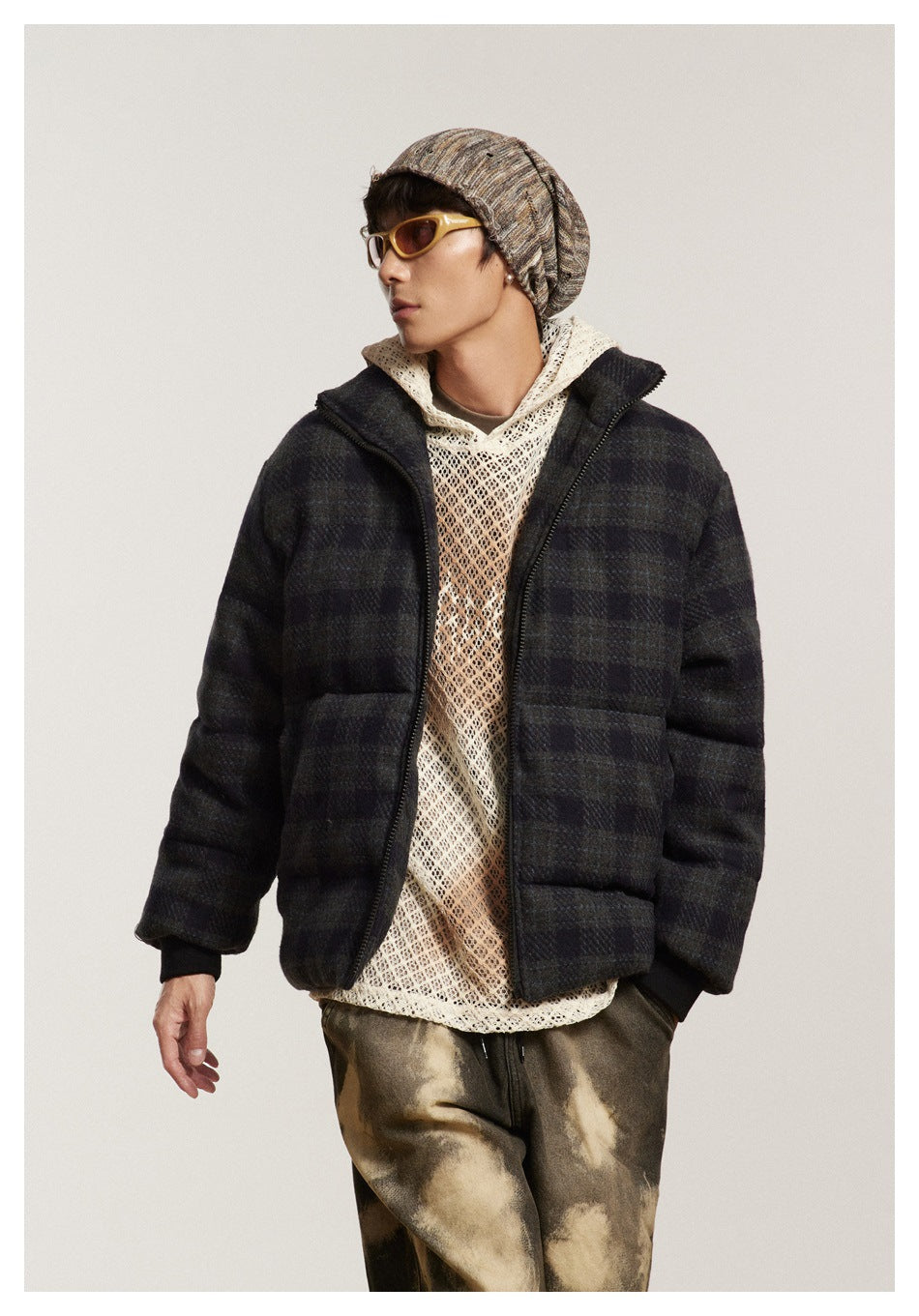 Model wearing an open dark plaid puffer jacket over a cream open-knit hoodie, with mottled brown pants.