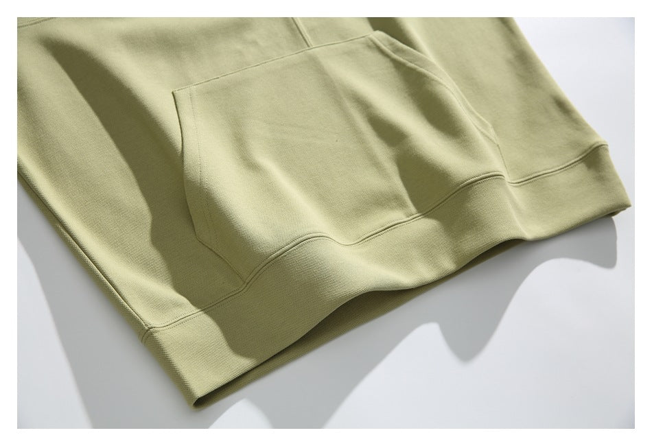 Close-up of the muted olive hoodie’s kangaroo pocket and ribbed bottom hem, laid flat.
