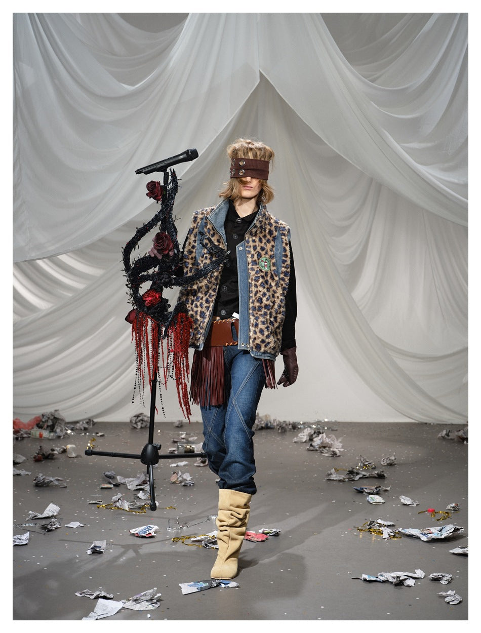 Model walks in a leopard-print, denim-trimmed fringed vest, dark jeans and tan boots beside a microphone stand.