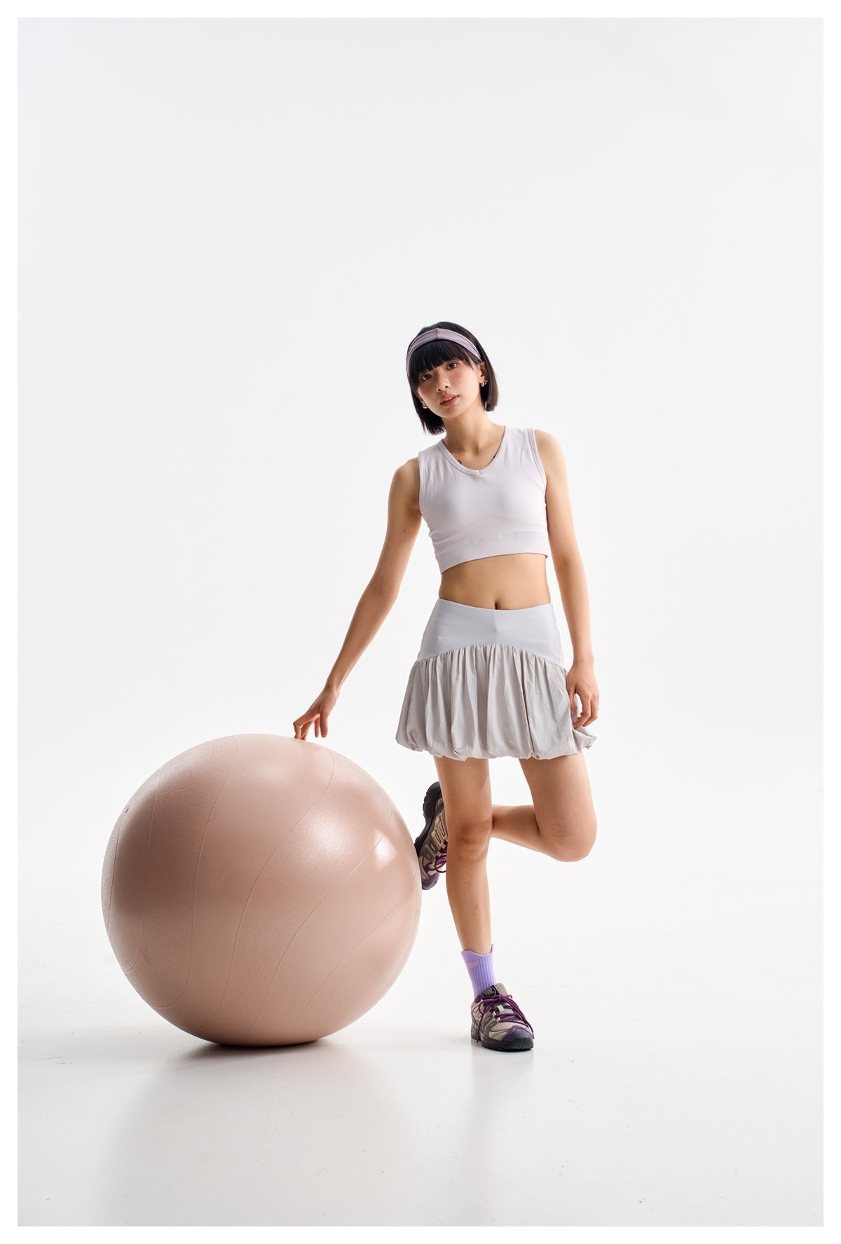 Model wearing a light gray bubble-hem athletic skirt with a white crop tank, posing beside a large exercise ball.