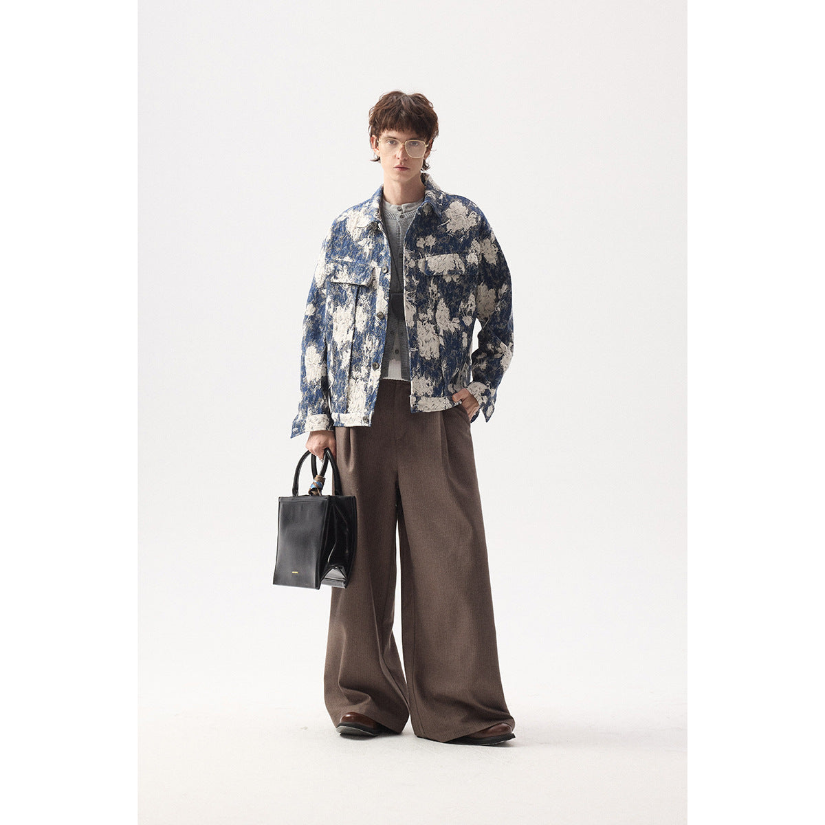 Full-length front view of a model wearing a blue and cream floral jacket with wide brown trousers and carrying a black bag.