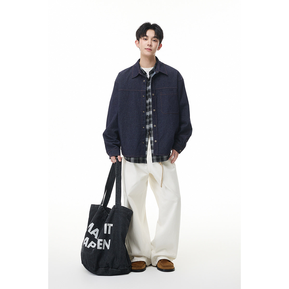 Full-length model wears a dark indigo jacket over a plaid shirt with loose white trousers and carries a printed black tote.