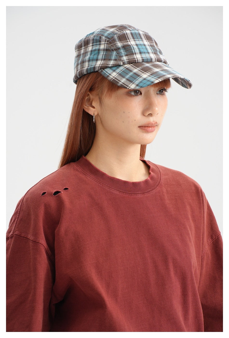 Close-up of a brick-red T-shirt's crew neck and distressed shoulder beside a blue-brown plaid cap.