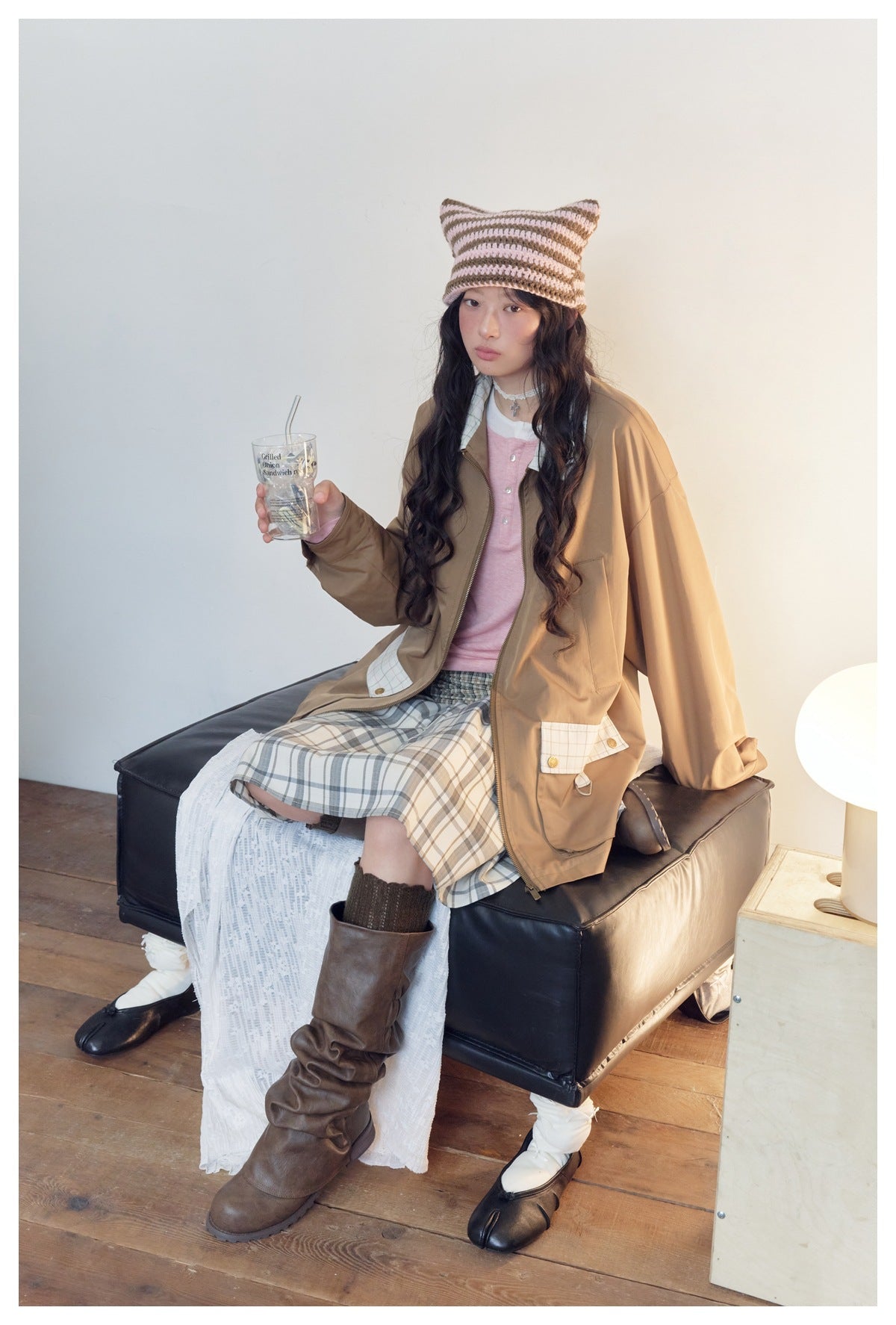 Model seated in a tan zip jacket with plaid collar and pocket flaps, pink top, plaid skirt, and striped knit cat-ear hat.