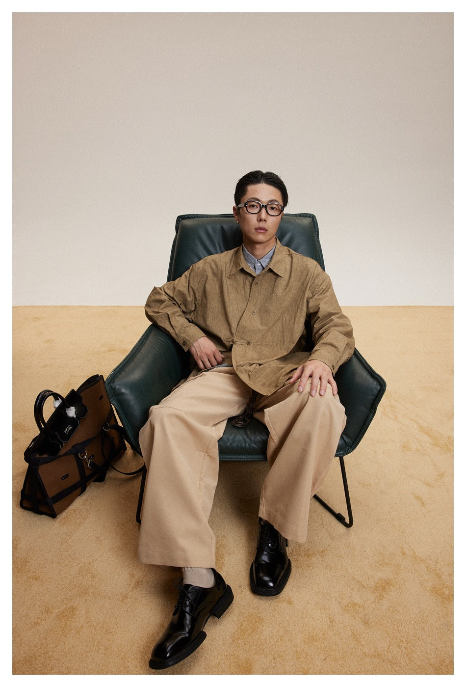 Seated model wearing a loose brown button-up shirt over a striped shirt and wide beige trousers.