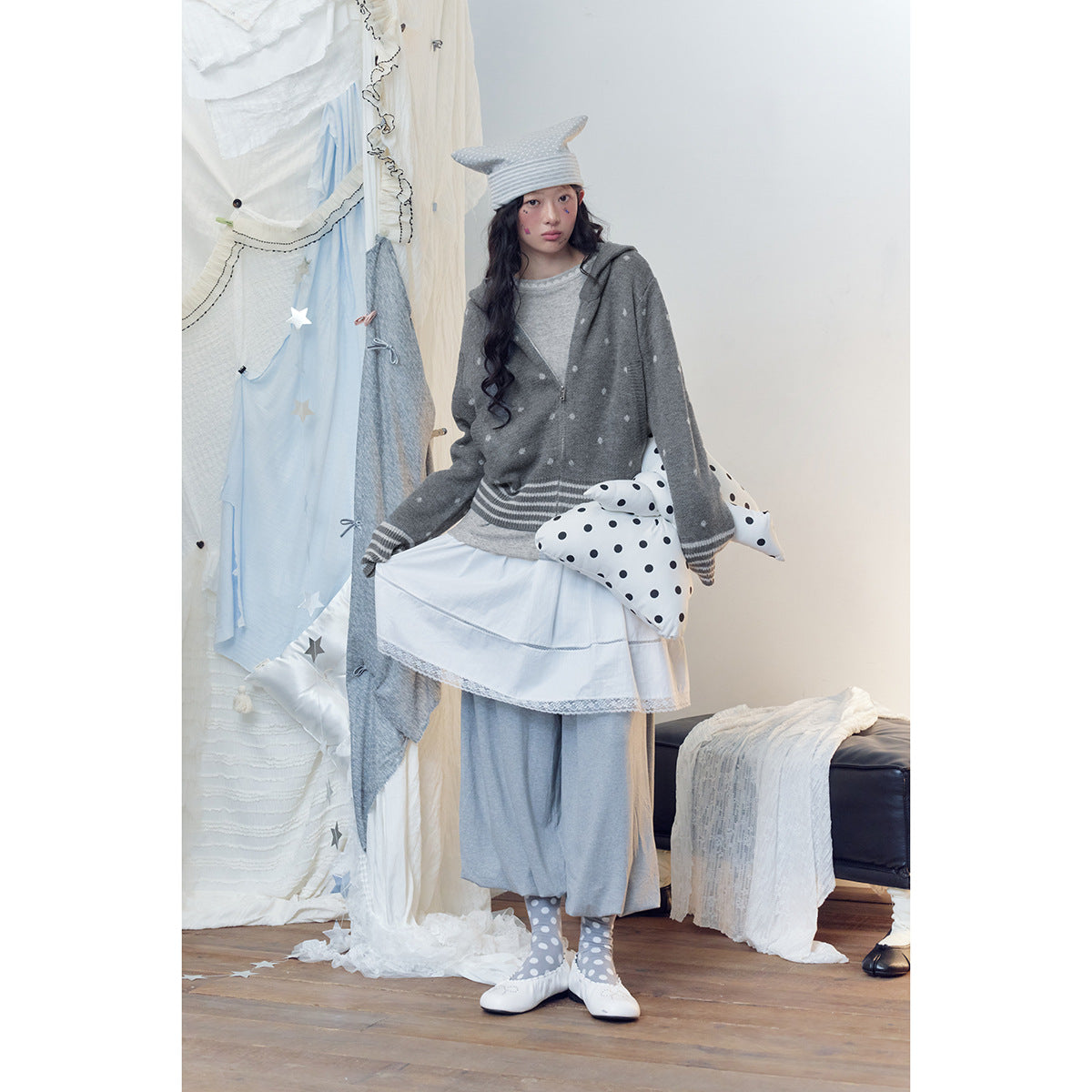 Full-length model wearing a gray spotted zip hoodie, layered white skirt, loose gray pants and polka-dot accessories.
