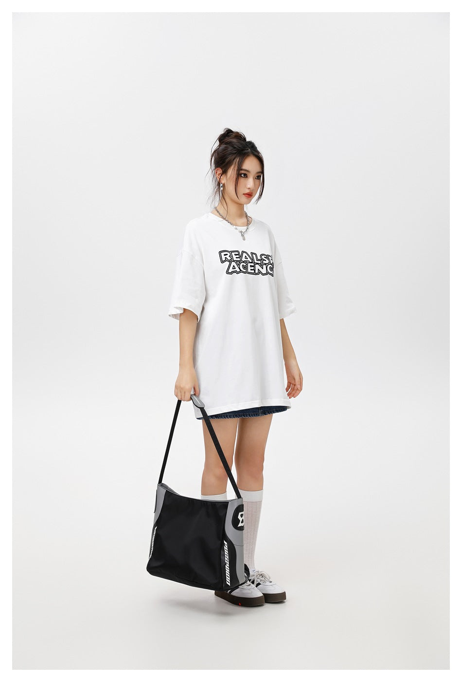 Model wearing an oversized white T-shirt with a lettered chest print, holding a black and gray bag