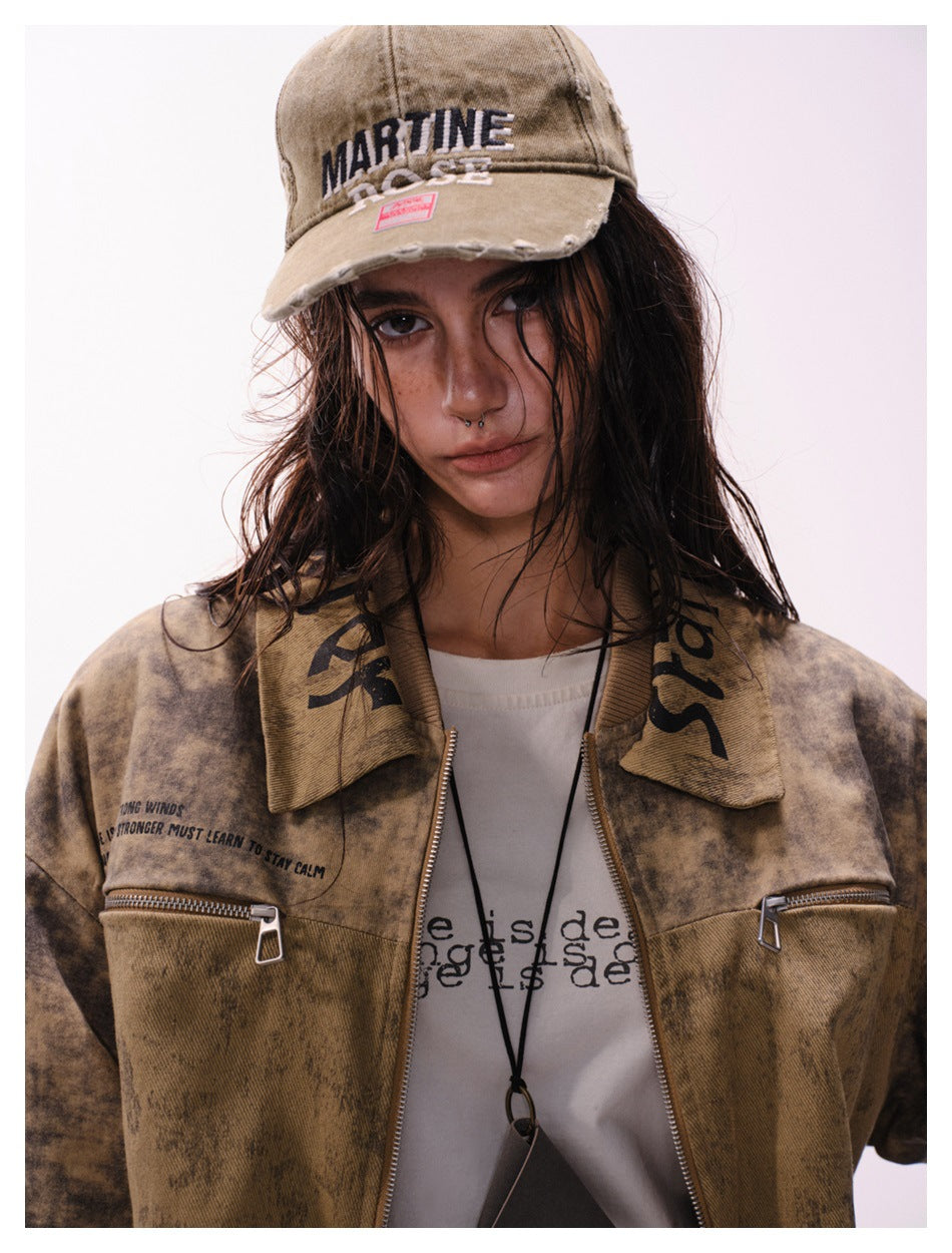 Close view of a model in a mottled brown jacket and distressed tan cap with embroidered lettering and chest zippers.
