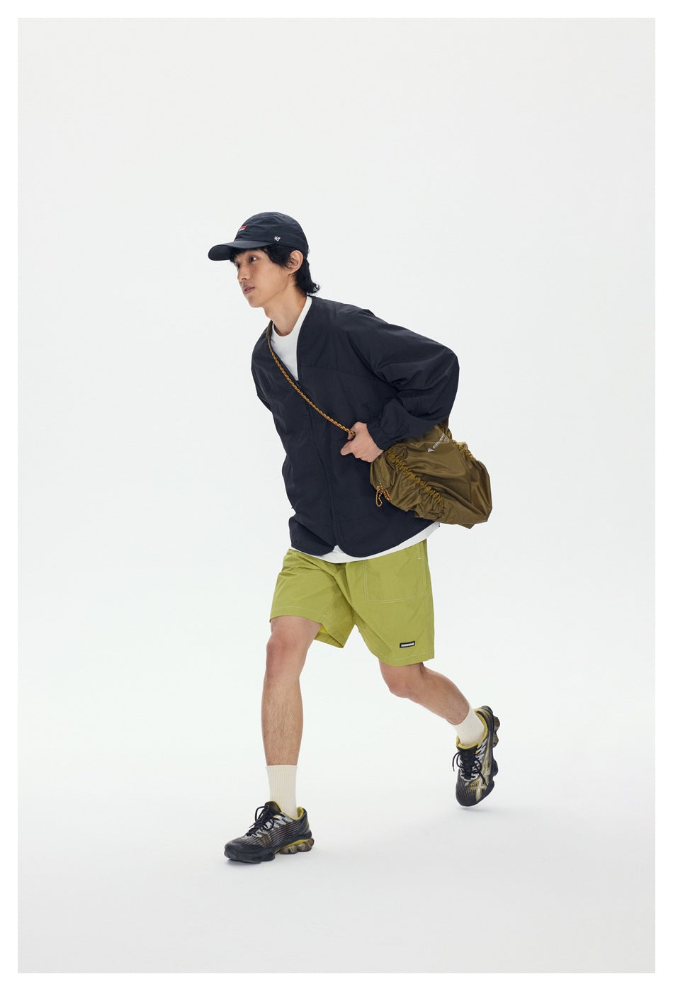Model in a black jacket and lime-green shorts, carrying a crossbody bag while walking.