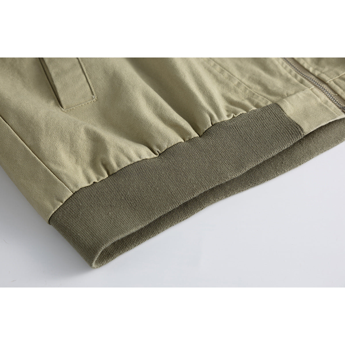 Close-up of a khaki jacket's gathered hem, dark olive ribbed waistband, pocket seam, and zipper edge.