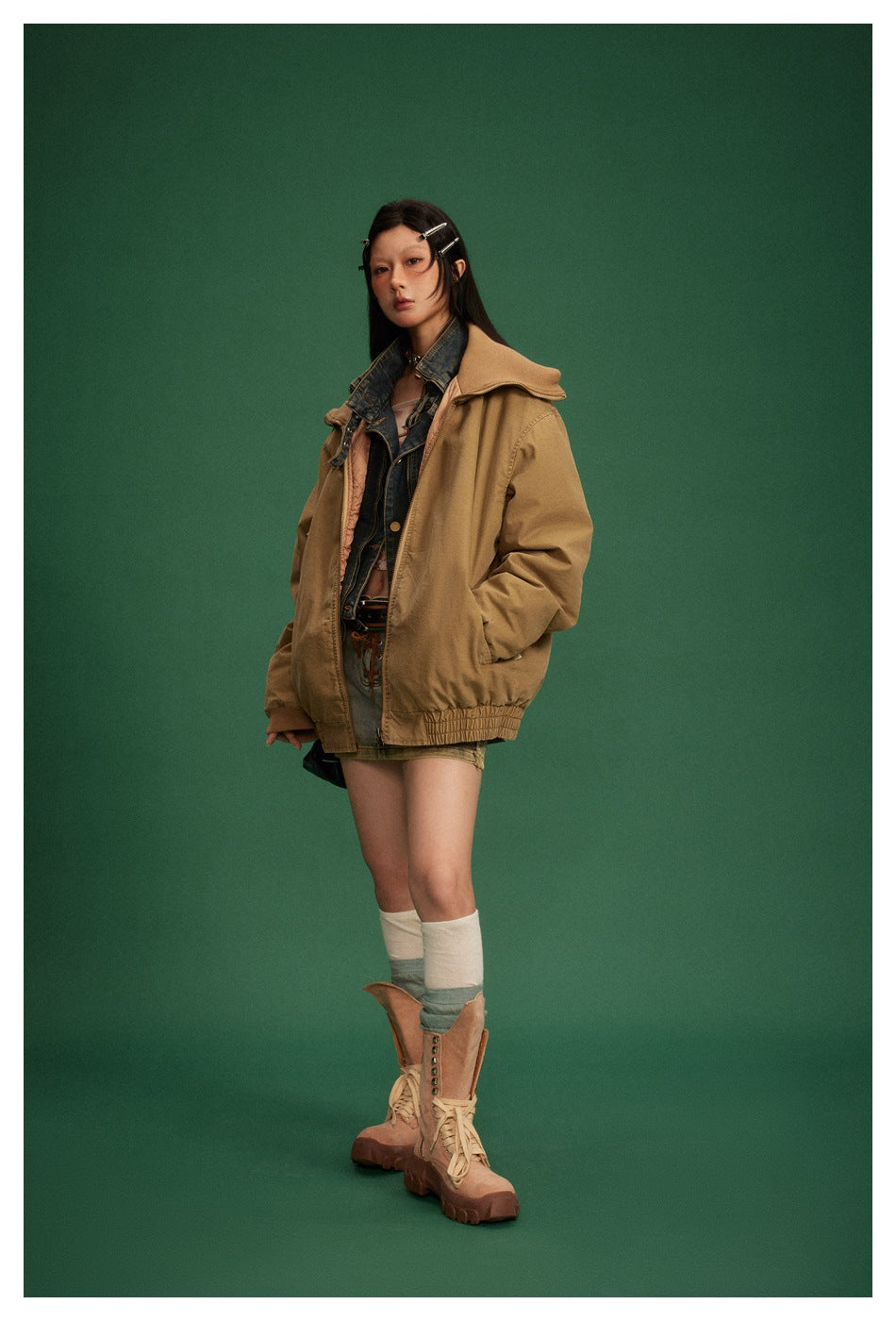 Model wearing a tan oversized jacket with wide collar over a denim vest, against a green backdrop