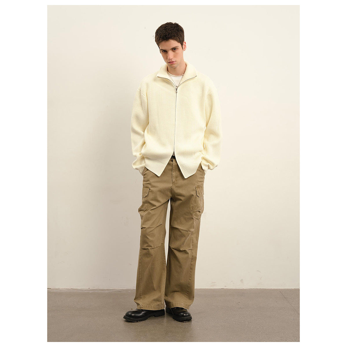 Front view of a model wearing an ivory ribbed zip-front sweater with a high collar and olive-brown cargo trousers.