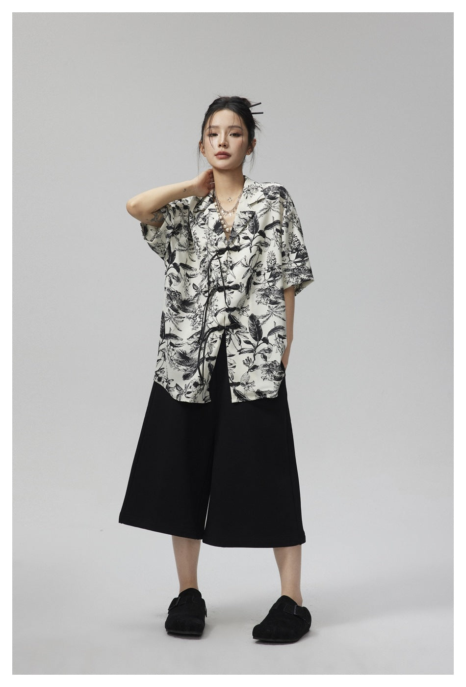 Model wearing an ivory short-sleeve shirt with black botanical print and frog closures, paired with black wide-leg cropped