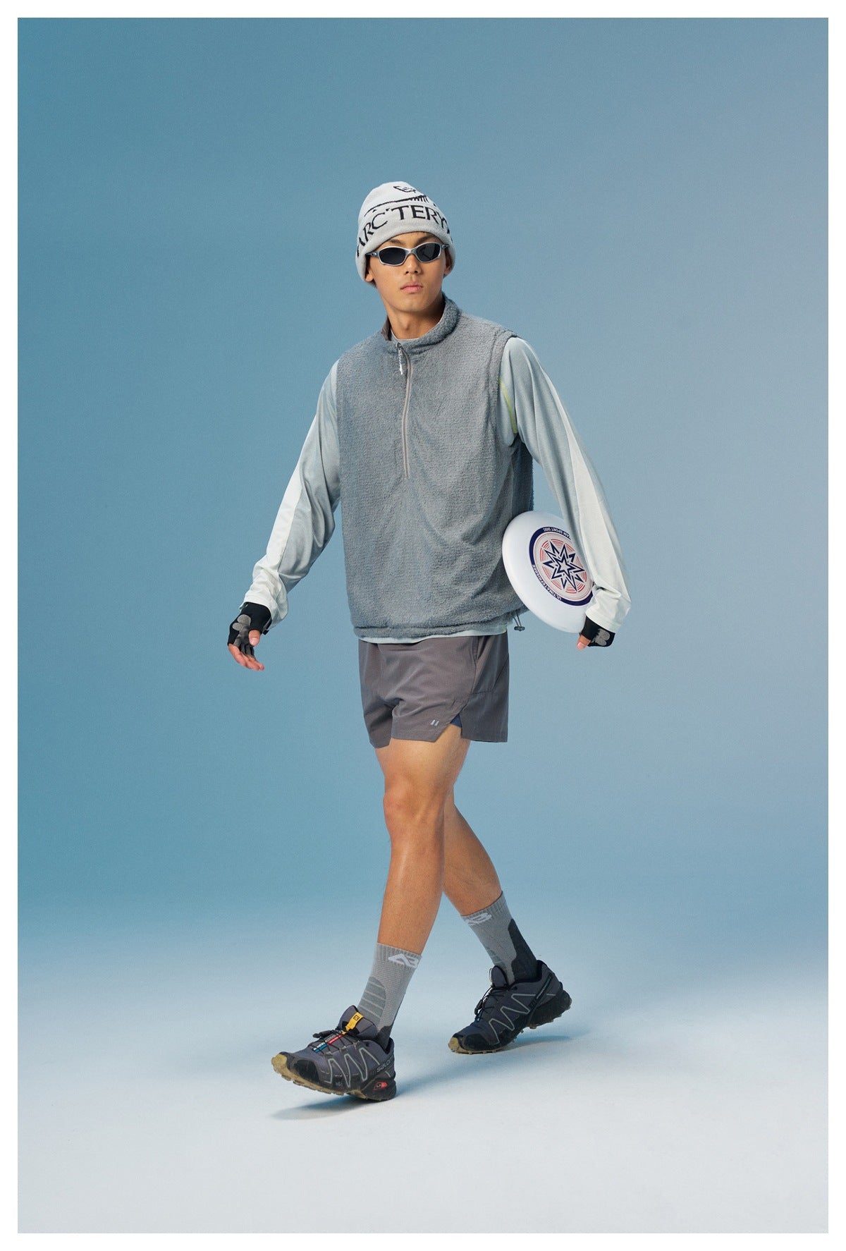 Model wearing a gray sleeveless zip vest over a pale long-sleeve top with gray shorts.