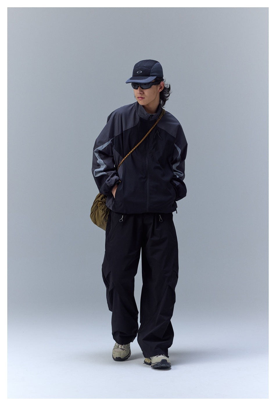 Model wearing a charcoal and black color-block zip jacket with loose black trousers, shown full length.