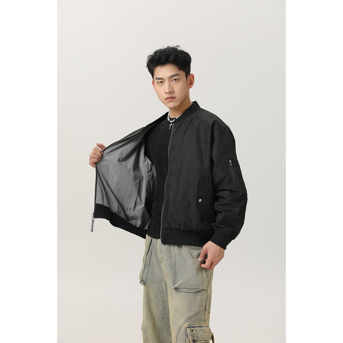 Model wearing a black bomber jacket, holding it open to show the gray lining, zip sleeve pocket and snap pocket.
