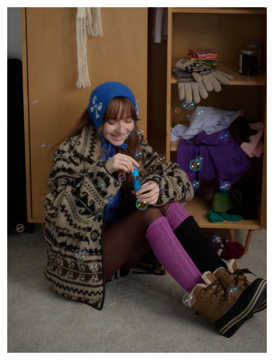 Seated model in a beige-and-black patterned zip jacket, blue bonnet, and pink and black leg warmers.