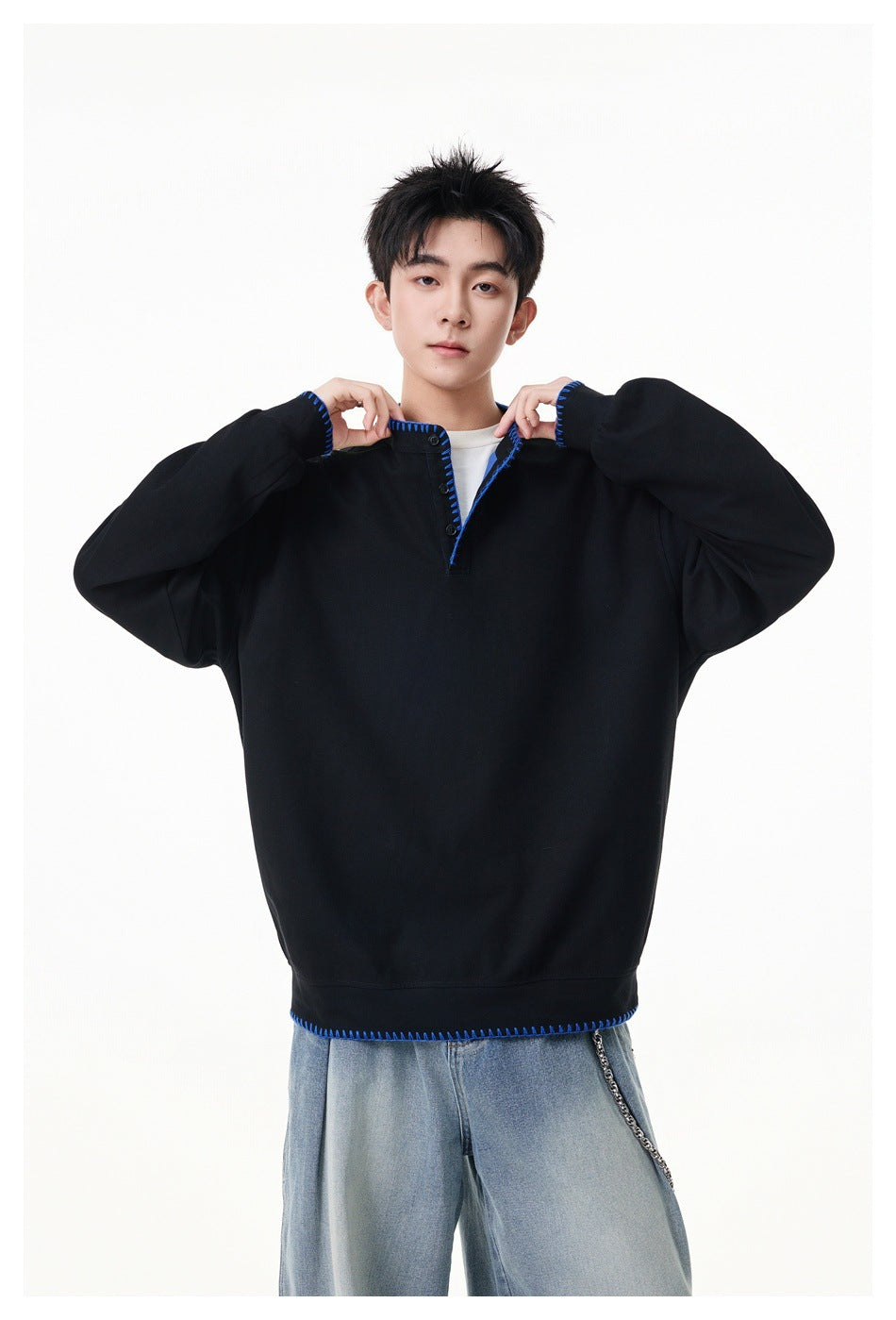 Model wearing a black oversized pullover with blue contrast stitching, a short button placket and ribbed hem.